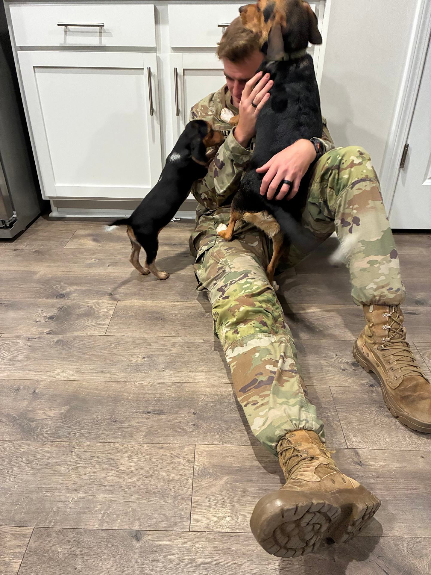A service member in uniform being welcomed home by two excited dogs